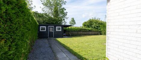 Ein kleiner, dunkel gefärbter Schuppen mit Tür und Fenstern, welcher in einem grasbewachsenen Hof mit Kiesweg steht.