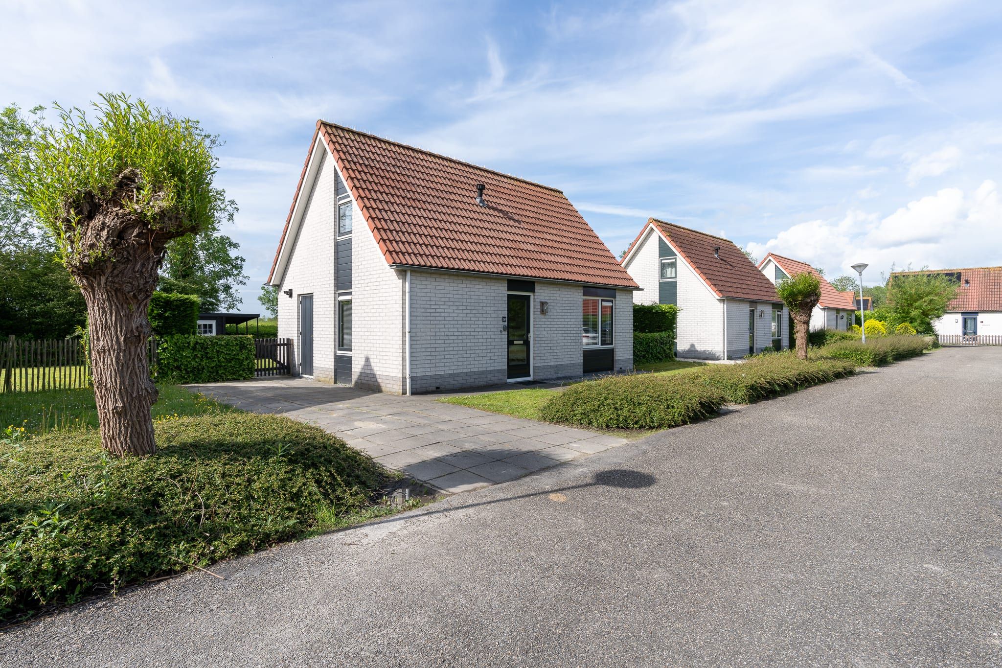 Une rangée de maisons en briques blanches, avec des toits recouverts de tuiles rouges, une allée pavée et une rue bordée d’arbres.