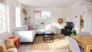 A modern living room with a sectional sofa, a round coffee table, a dining table with chairs, a television, and a large window with blinds.