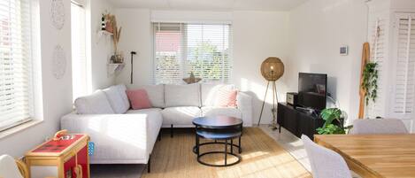 A modern living room with a sectional sofa, a round coffee table, a dining table with chairs, a television, and a large window with blinds.