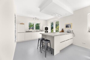 A modern kitchen with white cabinetry, a central island with stools, a built-in oven, and a window overlooking greenery.