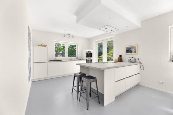 Una cocina moderna con armarios blancos, una isla central con taburetes, horno empotrado y ventana con vistas a la vegetación.