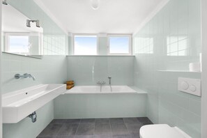 Un baño moderno con bañera, lavamanos e inodoro. Hay dos ventanas que dejan entrar luz natural.
