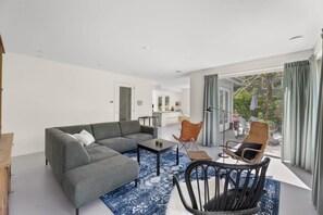 A modern living room with a grey sectional sofa, a small coffee table, and wicker chairs. A kitchen is visible in the background.