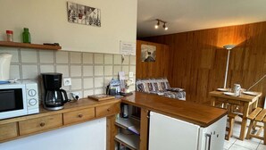 Cocina compacta con microondas, cafetera y refrigerador pequeño. Hay un mostrador de madera, una mesa de comedor de madera con sillas y un sofá en la zona de estar contigua.