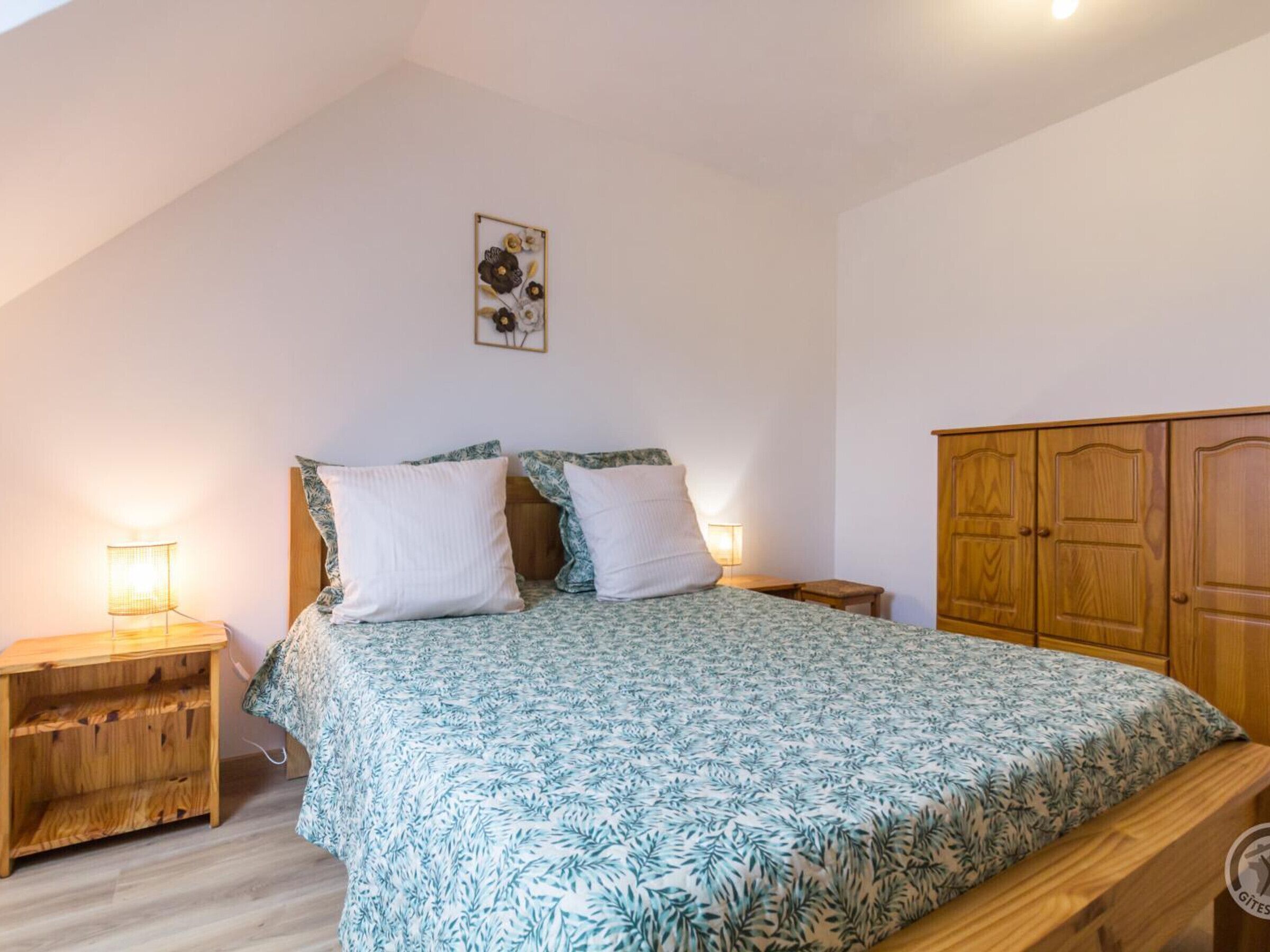 A bedroom with a bed, two bedside tables, a nightstand, a wooden wardrobe, and a wall-mounted artwork.