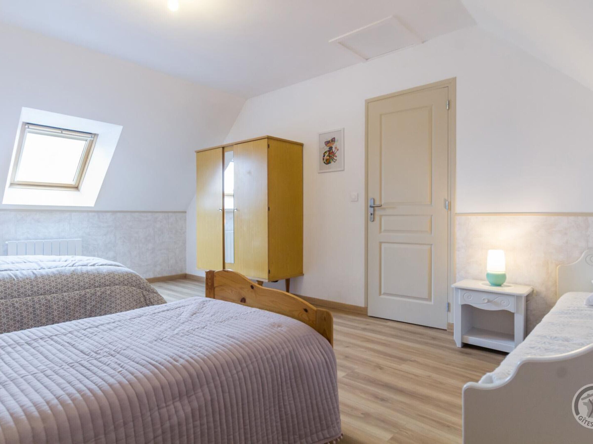 A room with two beds, a wardrobe, a door, a small bedside table with a lamp, and a window with a view.
