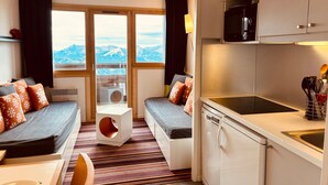 Una habitación de hotel compacta con sofá cama, una pequeña mesa de comedor, una zona de cocina con microondas y vistas a montañas nevadas a través de grandes ventanales.