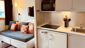Una kitchenette compacta con microondas, fregadero y lavavajillas. Un pequeño rincón de descanso con sofá y almohadas se encuentra junto a la cocina.