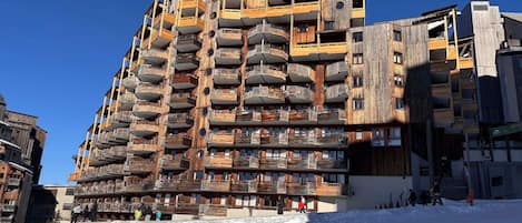 Un immeuble à plusieurs étages doté de balcons, une piste de ski au premier plan et un ciel bleu dégagé.