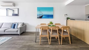 Ett modernt vardagsrum med en grå soffa, ett matbord i trä med stolar, en köksdel med mikrovågsugn och ett stort fönster med utsikt över havet.