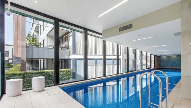 En inomhuspool med väggar av genomskinligt glas, en metallstege och två vita, cylindriska pallar.