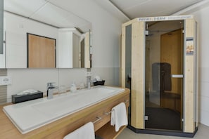 A modern bathroom with a large wooden vanity, a glass shower enclosure, and a visible sign for 'PHYSIO THERM Infrarotkabinen'.