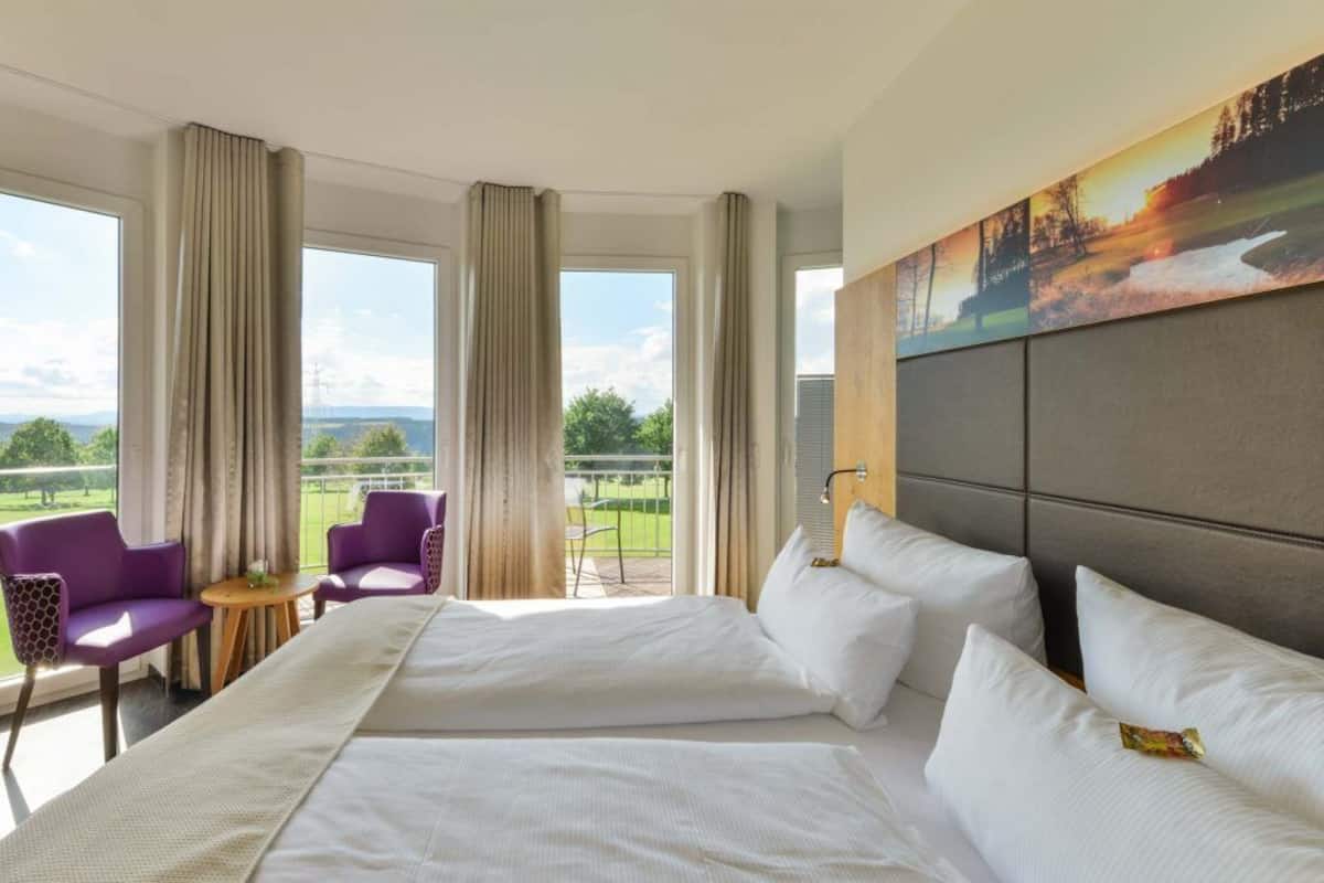 Ein Hotelzimmer mit einem großen Bett, zwei violetten Sessel, einem kleinen Tisch und einem Balkon mit Blick auf die Landschaft.