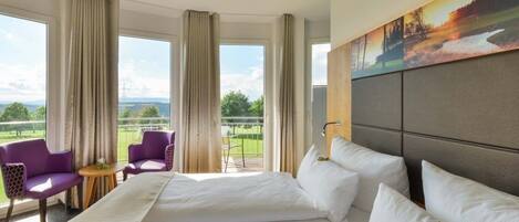 A hotel room with a large bed, two purple armchairs, a small table, and a balcony with a view of the countryside.