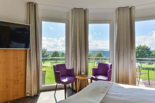 Una habitación de hotel con una cama grande, dos sillas moradas, una mesita y un televisor.