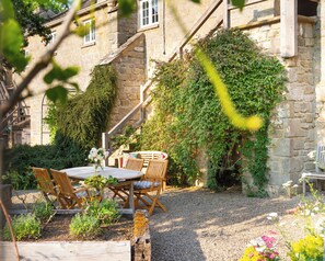A stone building with a wooden staircase, a round table with chairs, and a garden area with gravel and plants. - The Coach House at Jervaulx (Ripon)