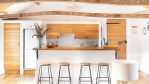 A modern kitchen with wooden cabinets, a central island with stools, and a dining area with a table and chairs. - The Coach House at Jervaulx (Ripon)