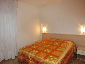 Un lit simple avec une couvre-lit à motifs, deux lampes de chevet, une table de chevet blanche et une fenêtre avec des rideaux blancs.