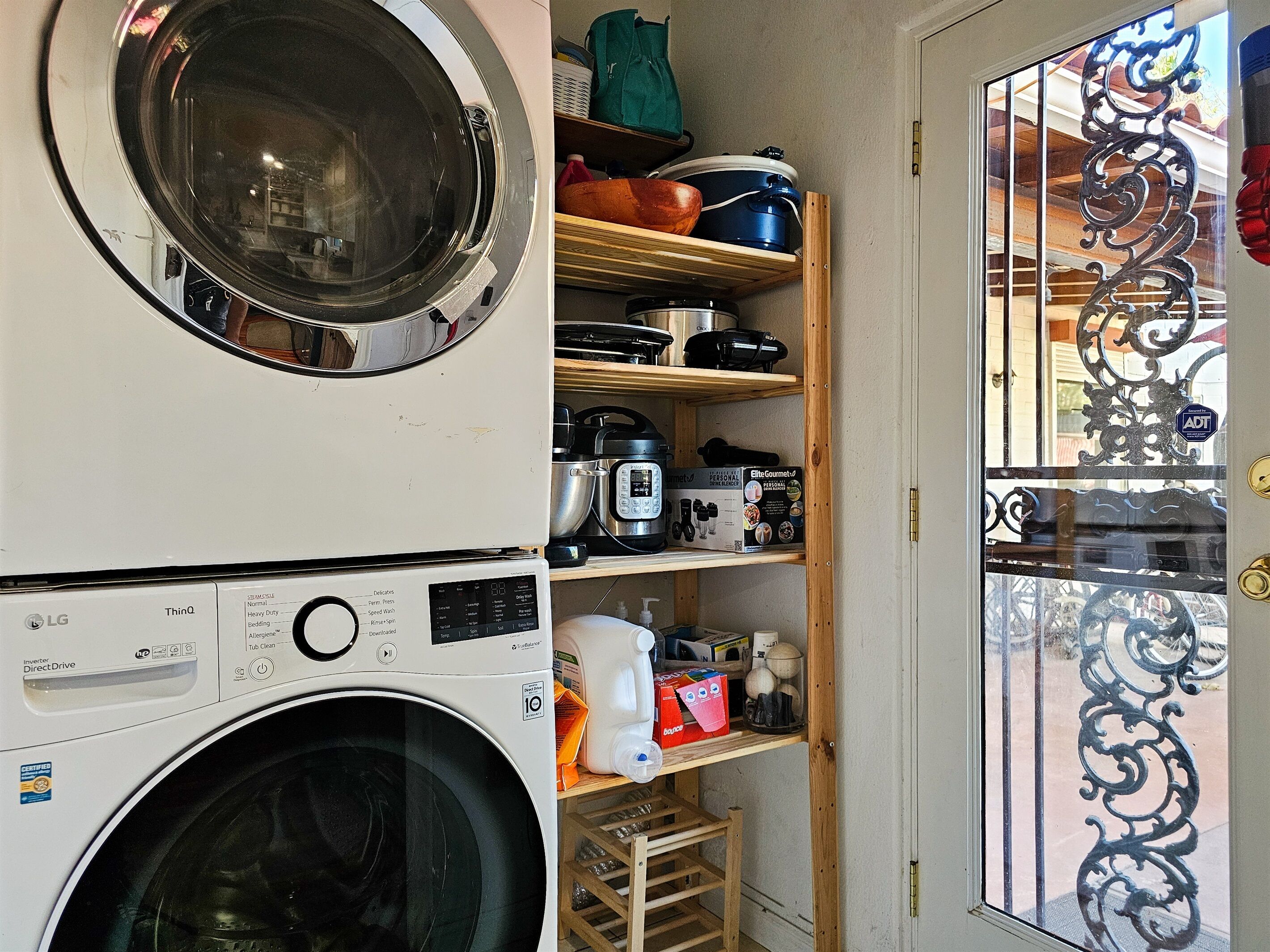 A laundry area with a front-loading washer and dryer, shelves with kitchen appliances, and a decorative metal gate.