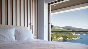 Une chambre avec un grand lit, des lattes en bois sur la tête de lit et une vue sur un paysage côtier, visible à travers une porte ouverte.