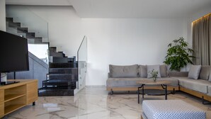 Un salon moderne avec un canapé, une table basse et un téléviseur. Un escalier avec une rampe en verre mène à l’étage supérieur.