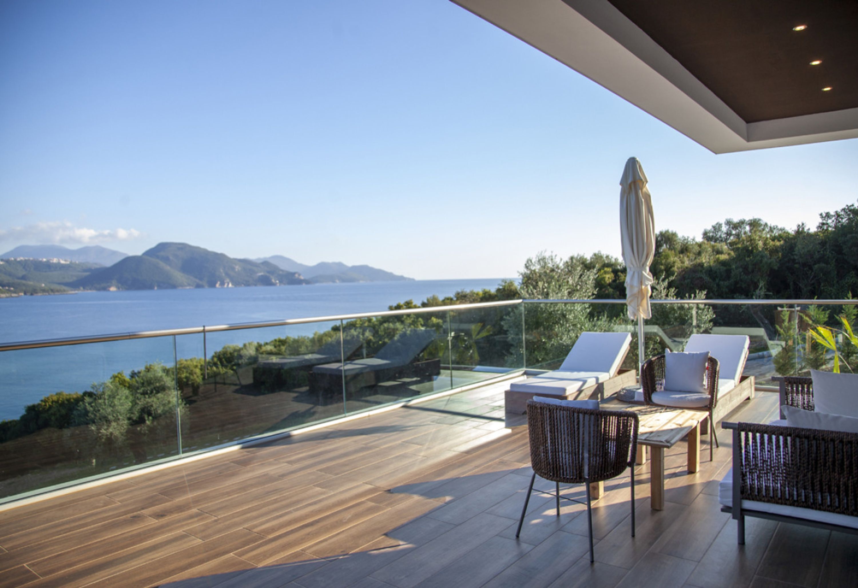 A modern outdoor terrace with a glass balustrade, wooden flooring, and seating areas overlooking a body of water and distant mountains.