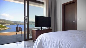 A hotel room with a large bed, a TV on a wooden dresser, and a sliding glass door leading to a balcony with a view of the sea. - Zavia Alpha in Syvota (Igoumenitsa)