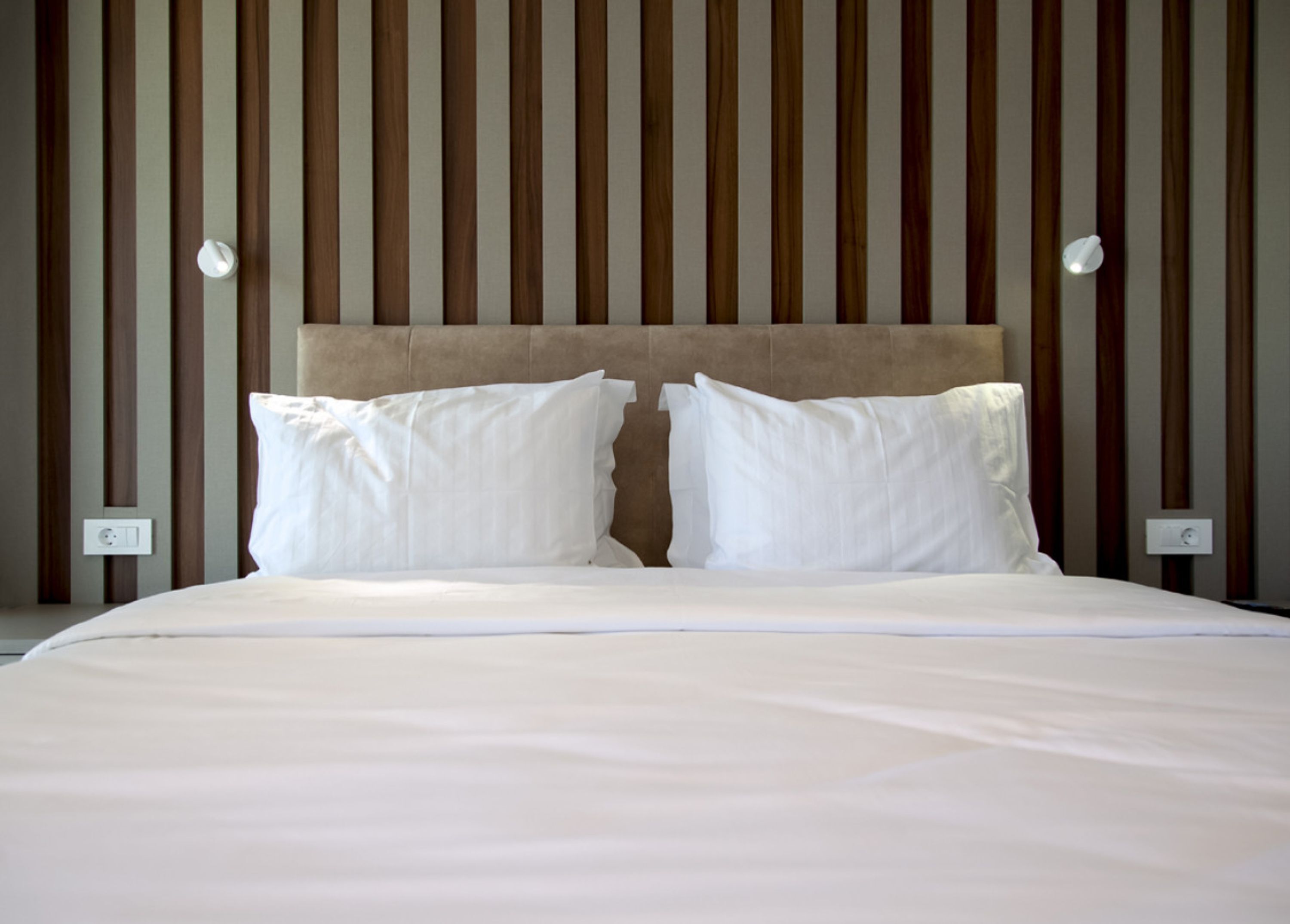 Un lit avec du linge de lit blanc et deux oreillers, placé contre un mur orné de panneaux de bois verticaux et agrémenté de deux appliques murales.