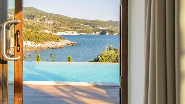 A view from a room with a wooden floor and a glass door, overlooking a pool and a coastal landscape with hills and a body of water. - Zavia Alpha in Syvota (Igoumenitsa)