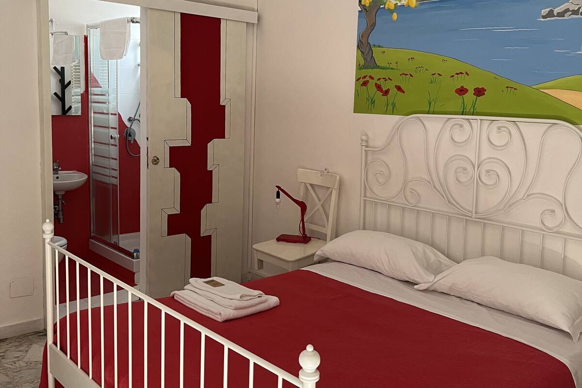Une chambre avec un lit blanc, une literie rouge, un luminaire suspendu rouge, un tableau représentant un paysage et une tête de lit blanche.