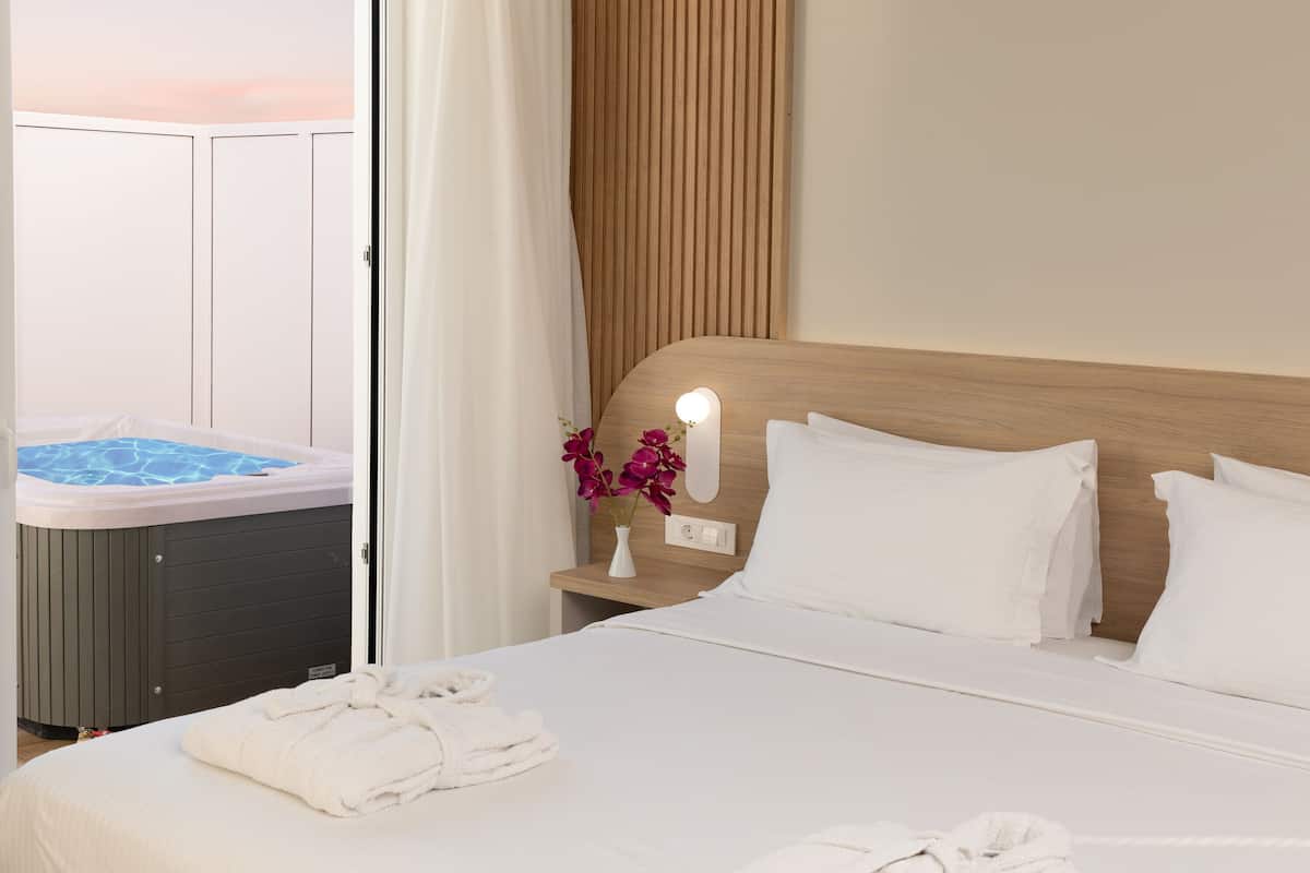 A hotel room with a bed, a nightstand with a lamp, a hot tub, and a view of the sky.