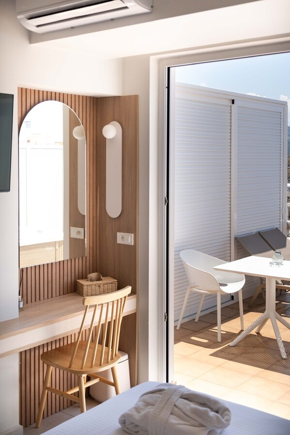 Una habitación moderna con un escritorio de madera, una silla, una cama con cubierta blanca y un ventanal que da a un espacio exterior con una mesa y sillas.