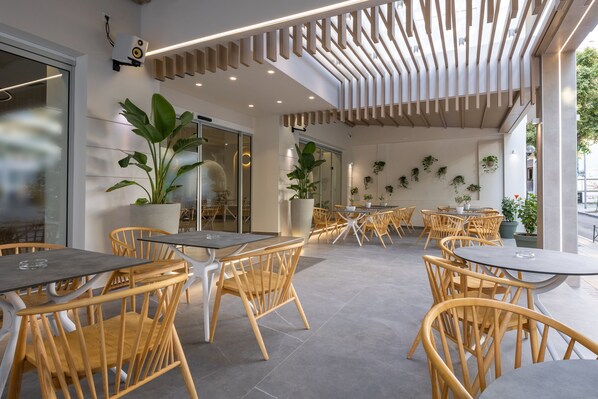 Un área de café al aire libre con sillas y mesas de madera, plantas en macetas y un diseño de techo moderno.