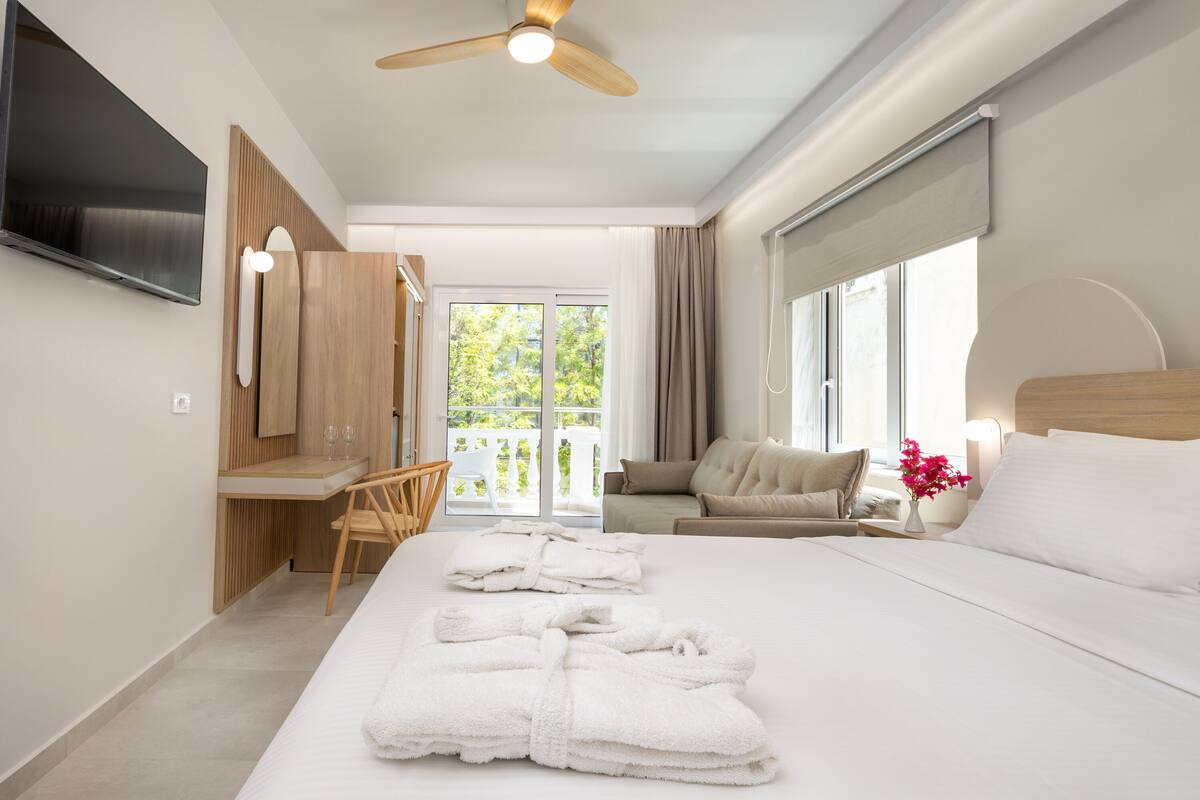 A modern hotel room with a large bed, a desk, a chair, a sofa, a TV, and a balcony with a view of greenery.