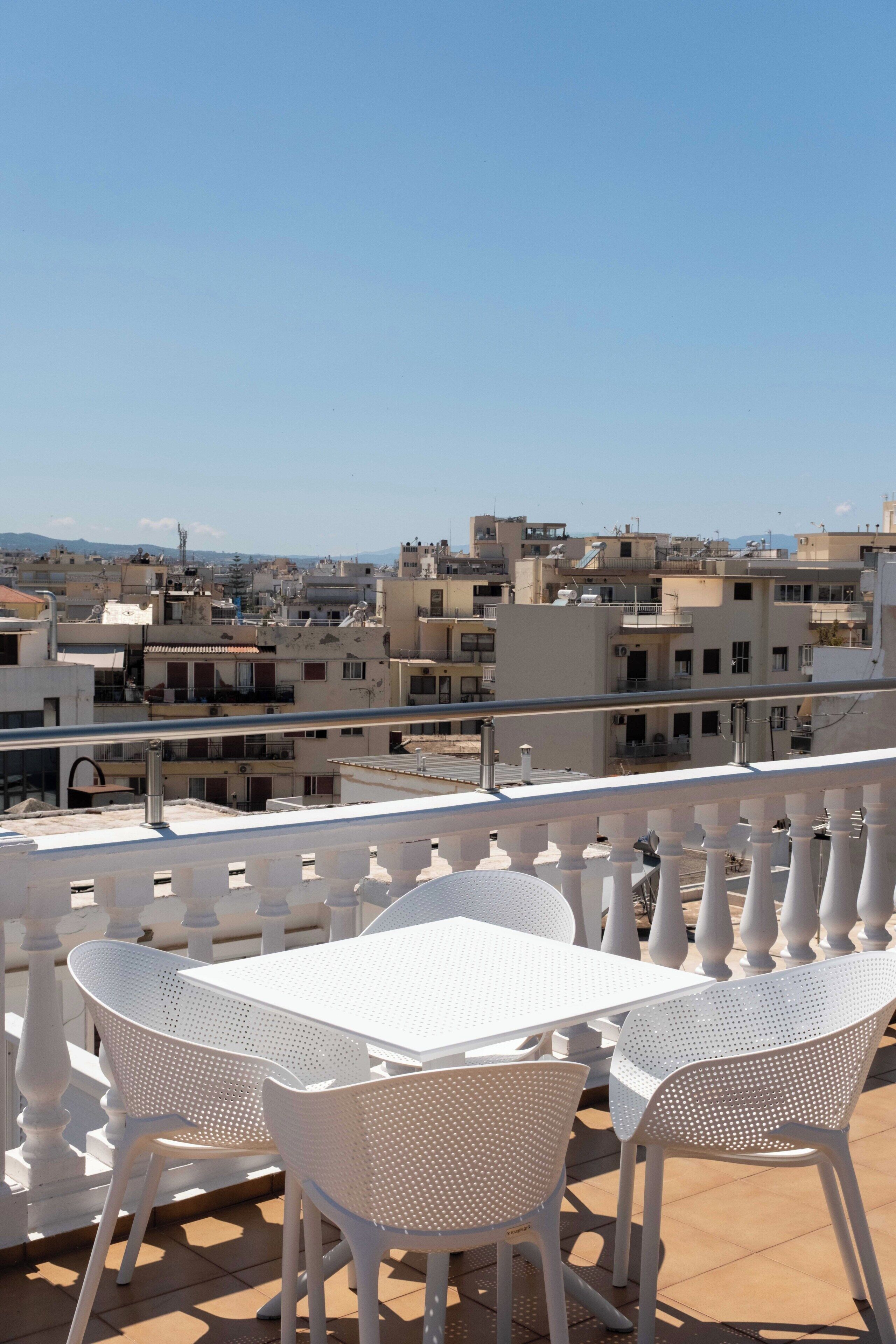 Una terraza con sillas y una mesa de metal blanco, con vistas a un paisaje urbano con edificios y cielo despejado.