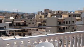 Una terraza con sillas y una mesa de metal blanco, con vistas a un paisaje urbano con edificios y cielo despejado.