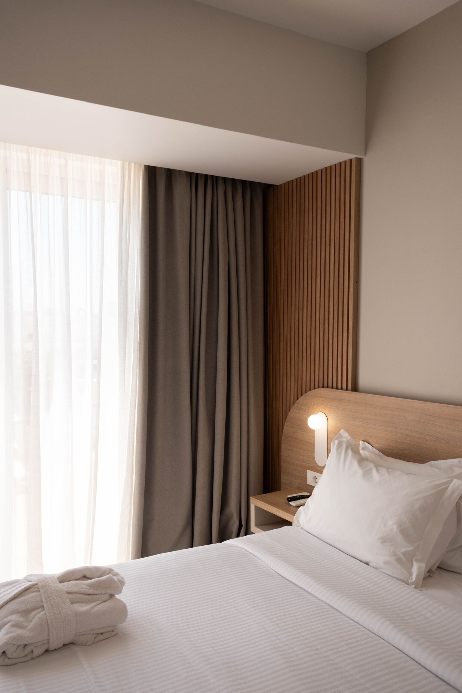 Une chambre d’hôtel avec un lit, une table de chevet, une lampe, une fenêtre avec des rideaux et un mur recouvert de panneaux de bois.
