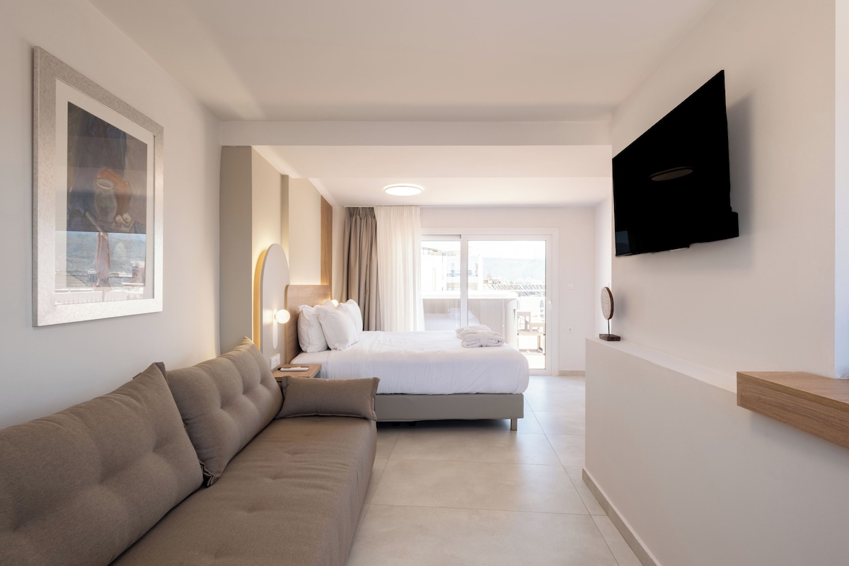 Una habitación de hotel moderna con cama, sofá, televisor montado en la pared y un ventanal grande.