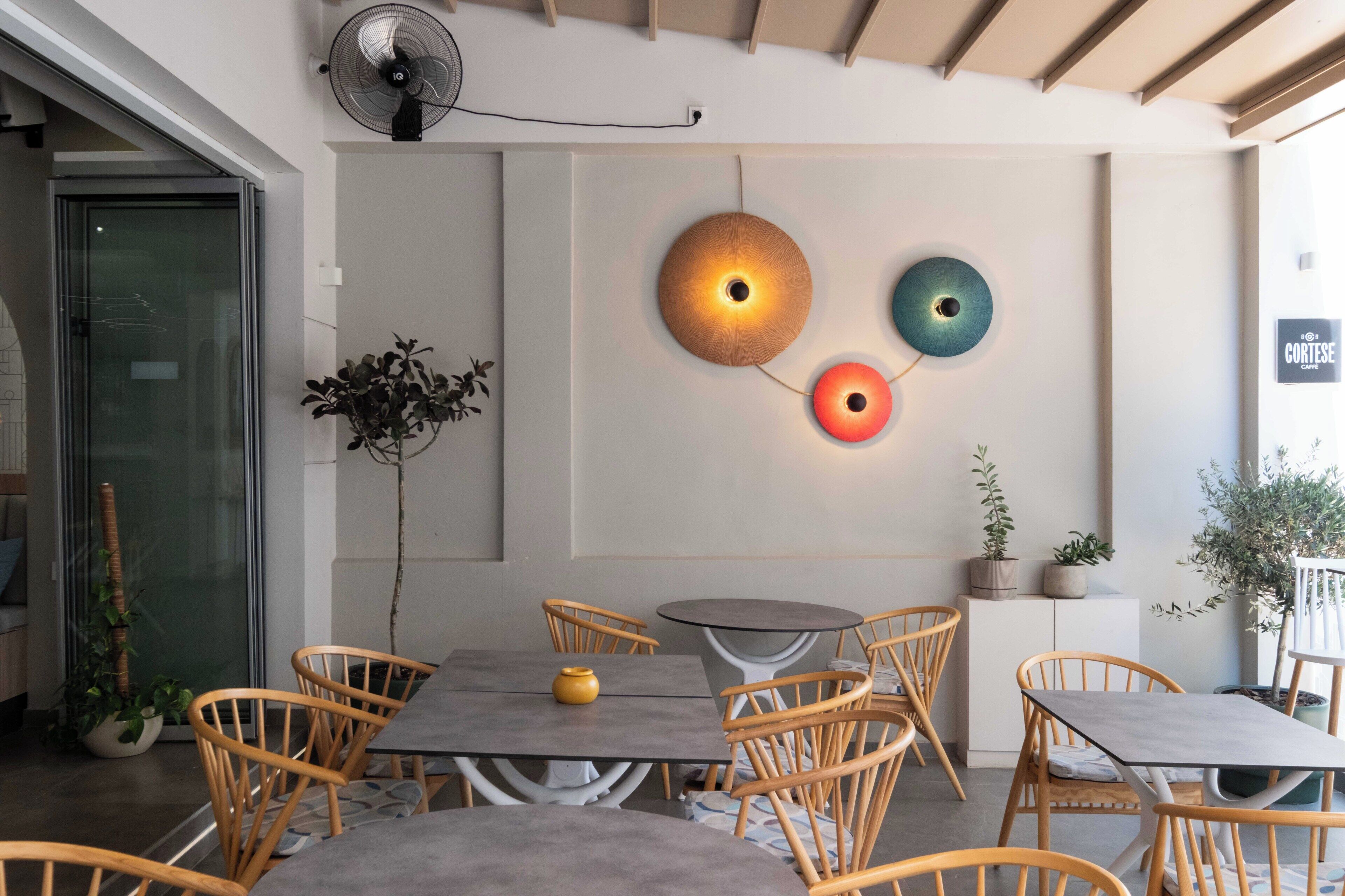 Un interior de cafetería moderno con mesas redondas, sillas de mimbre y luces decorativas en la pared.
