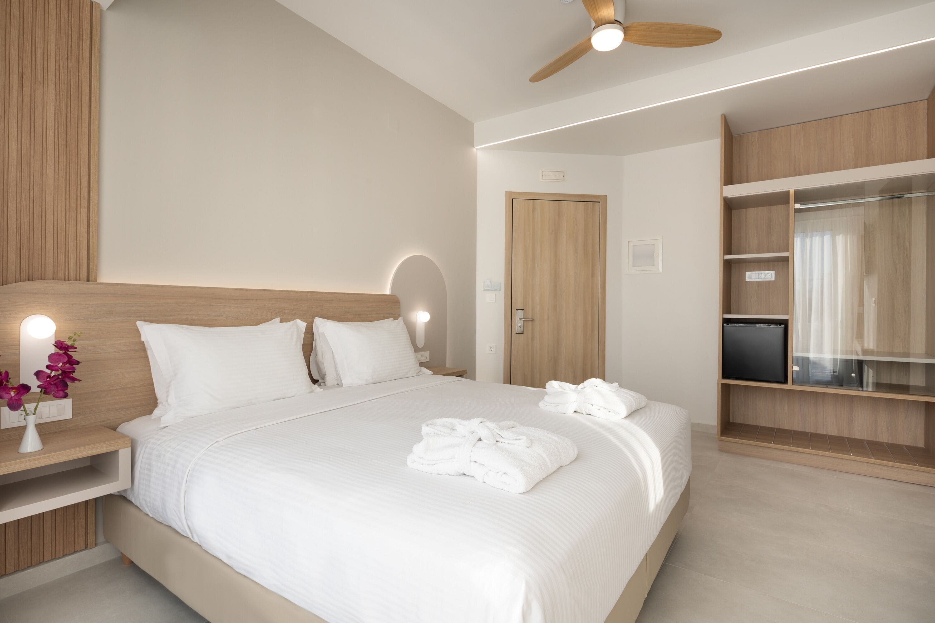 Une chambre d’hôtel moderne avec un grand lit, une tête de lit en bois, un ventilateur de plafond, une télévision et une armoire.