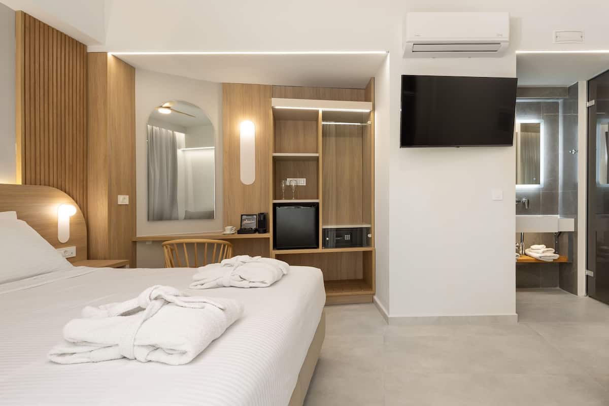 A modern hotel room with a bed, a desk with a chair, a TV, a wardrobe, and a bathroom with a mirror and sink.