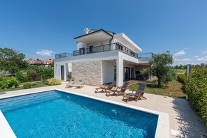 Ein modernes, zweistöckiges Haus mit Swimmingpool, Sitzgelegenheiten im Freien und einem Garten.