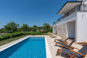 Ein Schwimmbecken mit Liegestühlen, einer Steinmauer und Blick auf Grünflächen.