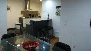 Et moderne køkken med en central ø, et spiseområde med et glasbord, og en bar med barstole.