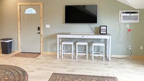 A room with a sloped ceiling, a white desk with two stools, a mounted TV, and an air conditioning unit. - Lariat Bunkhouse (Rapid City)