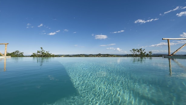 Ein klarer, blauer Infinity-Pool mit einer Holzterrasse auf einer Seite und Blick auf ferne Hügel und Bäume unter blauem Himmel.