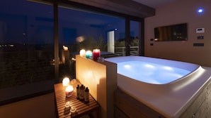 Ein modernes Badezimmer mit einer großen Badewanne, einem Holzbadezimmerhocker mit Kerzen und einem großen Fenster mit Blick auf eine nächtliche Skyline.