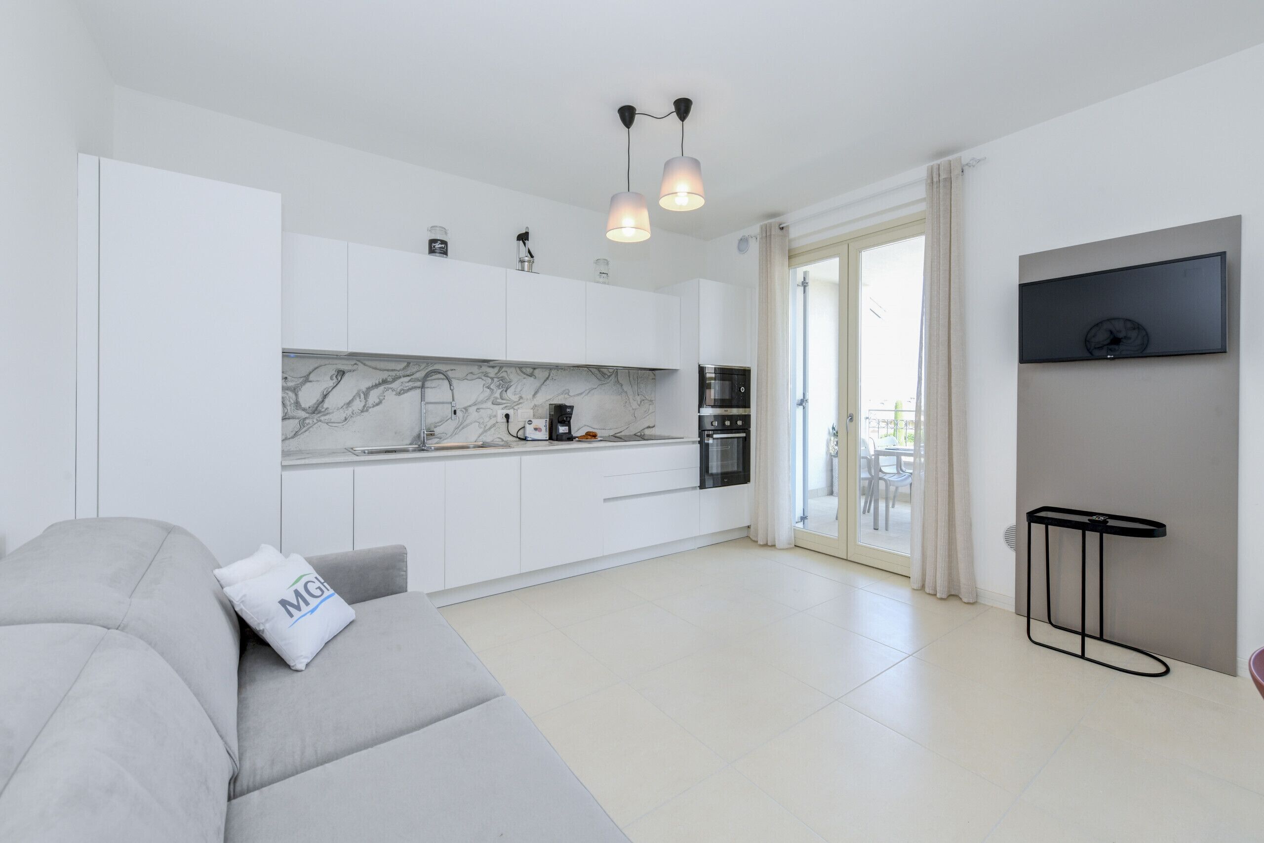 Eine moderne Küche mit weißen Schränken, einer Marmor-Spritzschutzwand und einer schwarzen Arbeitsplatte. Ein graues Sofa mit Kissen, ein schwarzer Beistelltisch und ein an der Wand befestigter Fernseher.