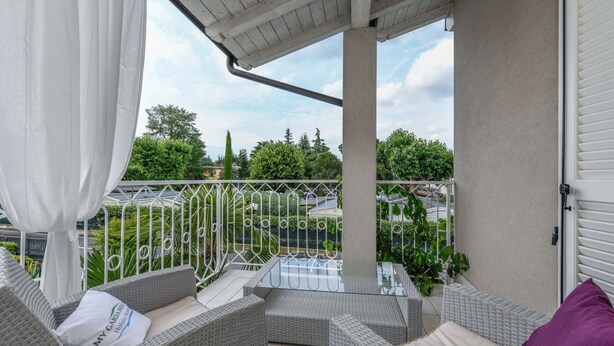 A balcony with wicker furniture, a glass table, and a view of greenery and buildings.